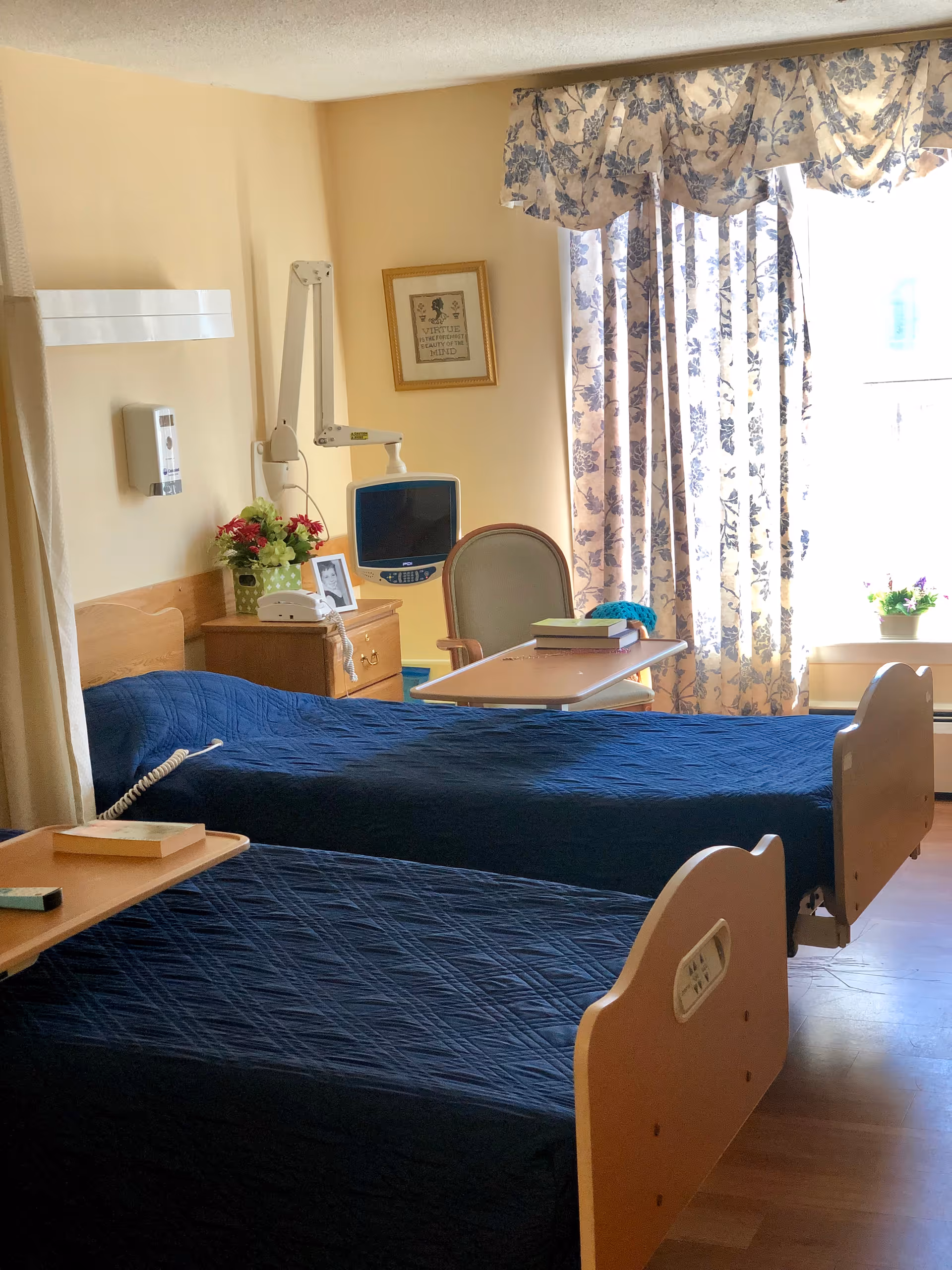 Sunlit room with two hospital-style beds covered in dark blue quilts, a bedside table and chair, a medical monitor, and floral curtains.