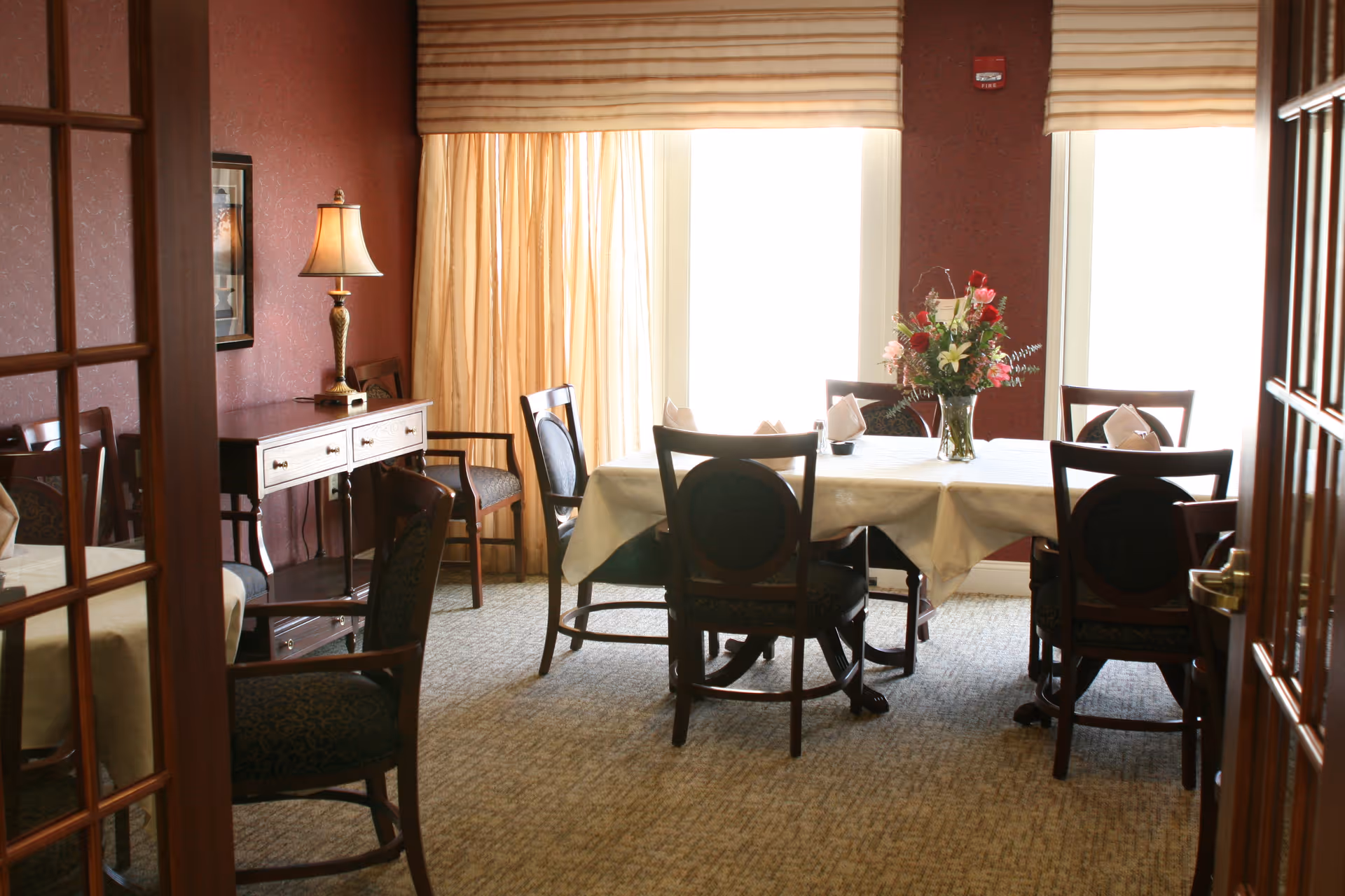 A dining room with a rectangular table covered with a white tablecloth, surrounded by six wooden chairs with cushioned seats. A vase with a colorful flower arrangement is placed in the center of the table. The room has carpeted flooring, maroon walls, two large windows with beige curtains, and a side table with a lamp and additional chairs.