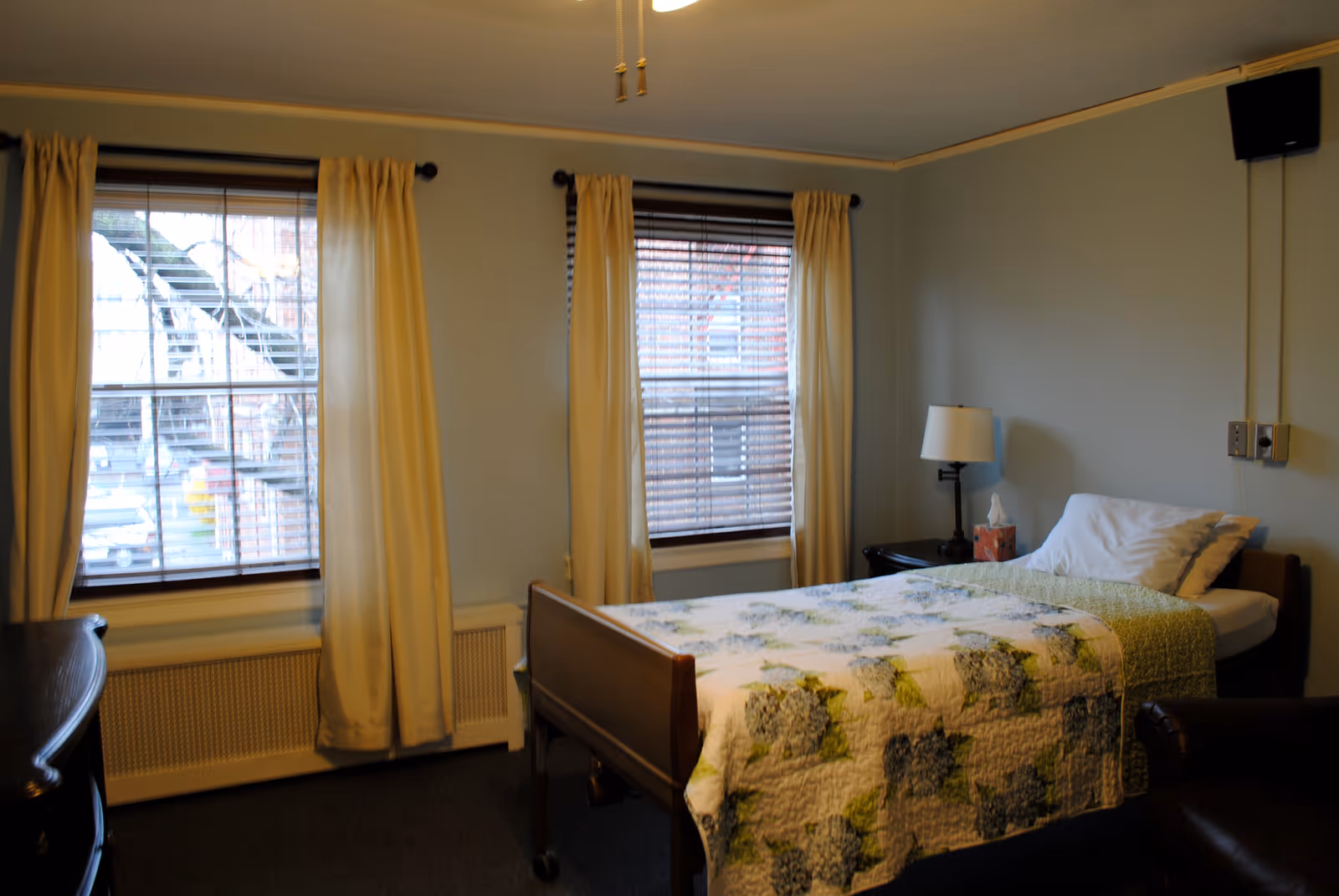 A simple bedroom with a single bed covered with a floral quilt, two windows with yellow curtains and blinds, a bedside table with a lamp and tissue box, and a ceiling fan with pull chains.