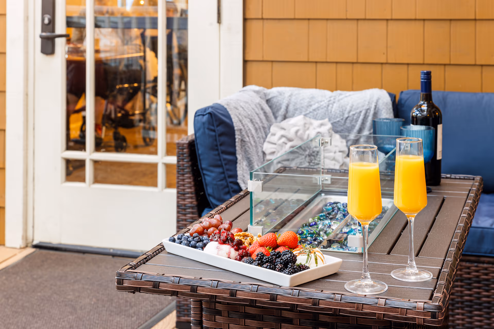 Outdoor patio seating area with a wicker table and blue cushioned chairs. On the table, there is a rectangular white plate with assorted fresh berries and grapes, two glasses filled with orange juice, a bottle of wine, and two blue cups. A gray blanket is draped over one of the chairs, and a glass door with white framing is visible in the background.