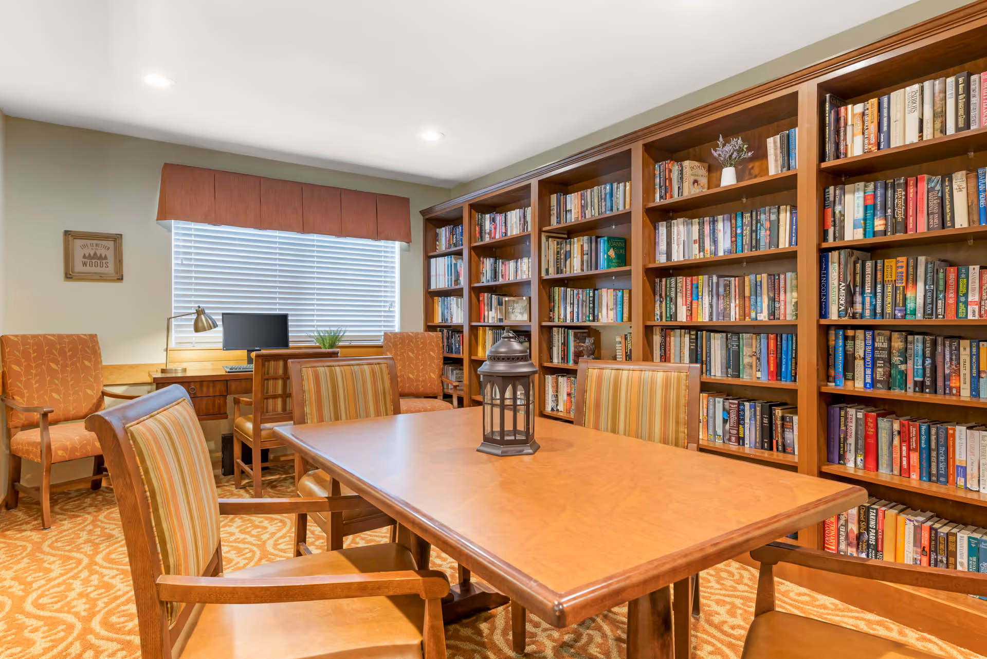 A cozy library room with wooden bookshelves filled with books along one wall. In the center, there is a wooden table with four striped cushioned chairs around it and a decorative lantern on top. In the background, there is a desk with a computer, a lamp, and two orange upholstered chairs. A window with blinds and a brown valance lets in natural light. The carpet has a patterned design in warm tones.