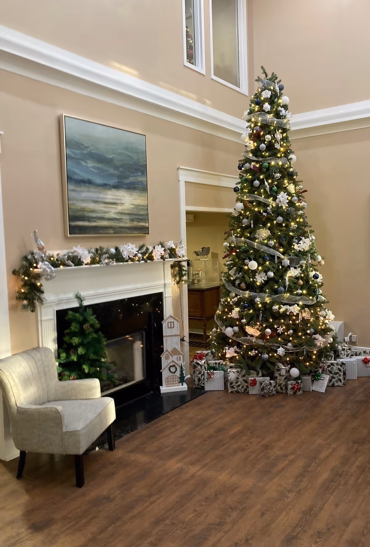 A decorated living room lobby with a tall Christmas tree, wrapped gifts, a fireplace mantel garland, and an upholstered chair.