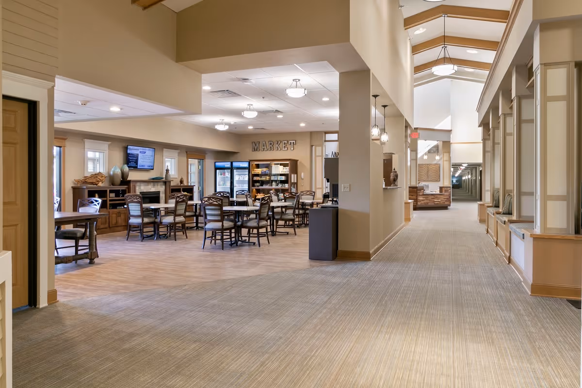 Interior view of a senior living facility showing a spacious hallway with built-in seating along the right wall and a dining area labeled 'MARKET' with tables and chairs on the left. The space is well-lit with ceiling lights and has a warm, neutral color palette with wood accents.