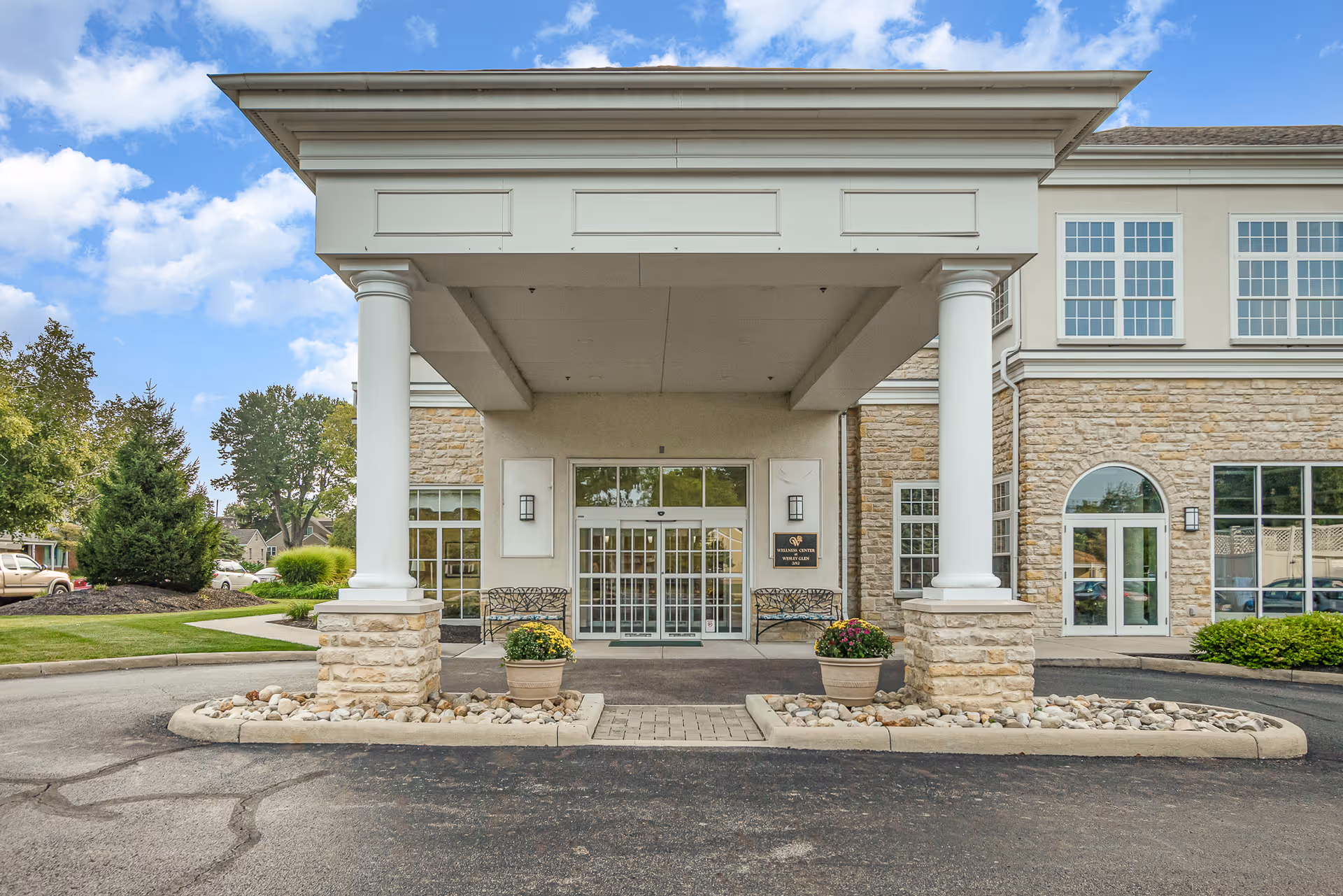 Front entrance of Wesley Glen with a covered portico supported by white columns, stone facade, glass doors, benches and potted plants.