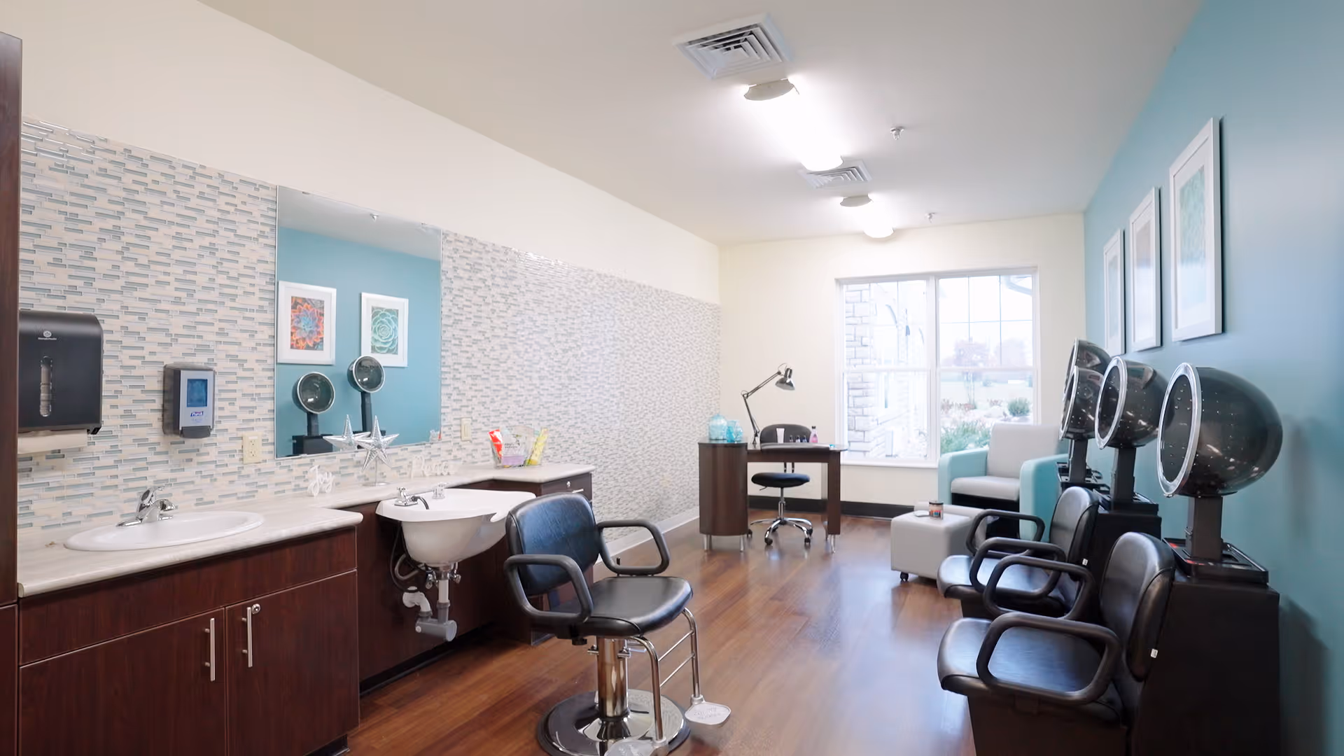 A bright and clean salon area in a senior living facility with three black salon chairs facing hair drying stations on the right. On the left, there is a countertop with a sink, a wash basin, and a large mirror above. The room has light blue and white walls, wooden flooring, and a window at the far end letting in natural light. A small desk with a chair and a light is positioned near the window.