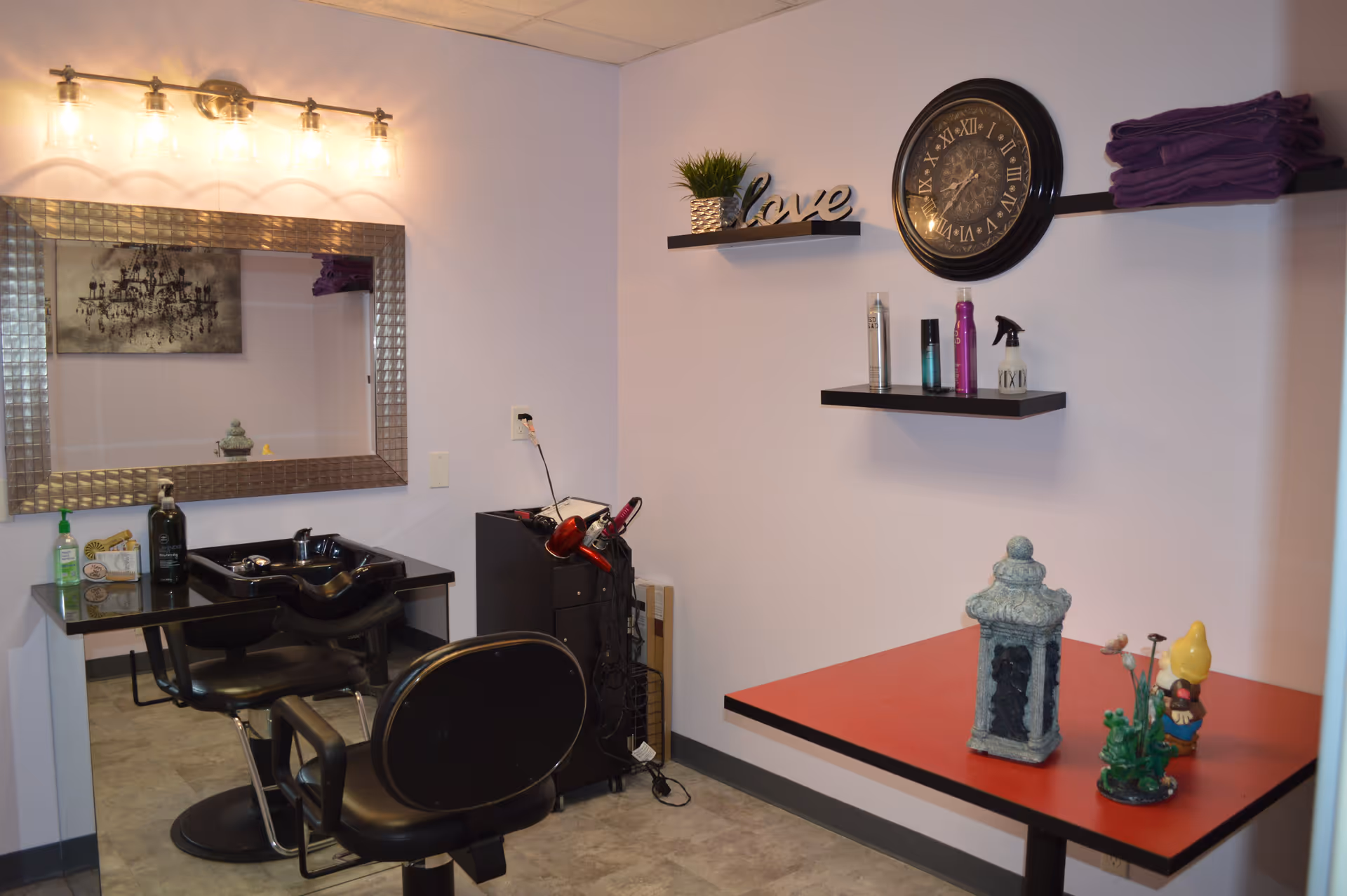 Interior of a salon area with two black salon chairs in front of a black sink and a large rectangular mirror with a metallic frame. Above the mirror is a light fixture with five exposed bulbs. On the right wall, there are two black floating shelves holding decorative items including a small plant, a clock with Roman numerals, hair products, and folded purple towels. A red table below the shelves holds decorative figurines and a small statue.