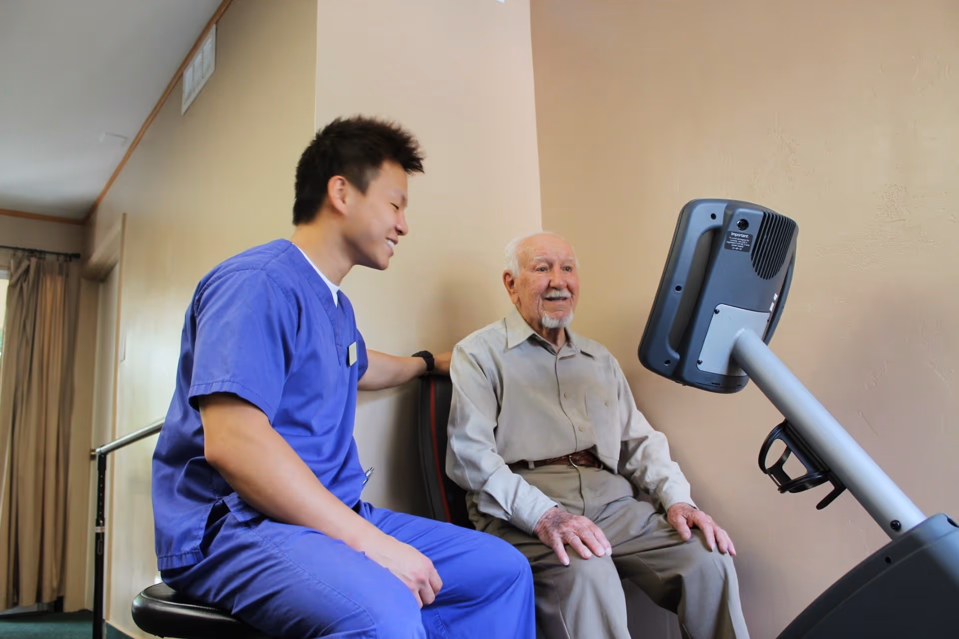 A young male caregiver in blue scrubs is sitting next to an elderly man who is seated on a chair in front of a medical or exercise device mounted on a stand, both smiling and interacting in a room with beige walls.