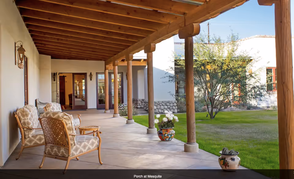 Covered porch area with wooden beams and columns, featuring cushioned chairs and small tables. The porch overlooks a green lawn with trees and decorative potted plants near the edge of the porch. The building has white walls and large windows and doors.