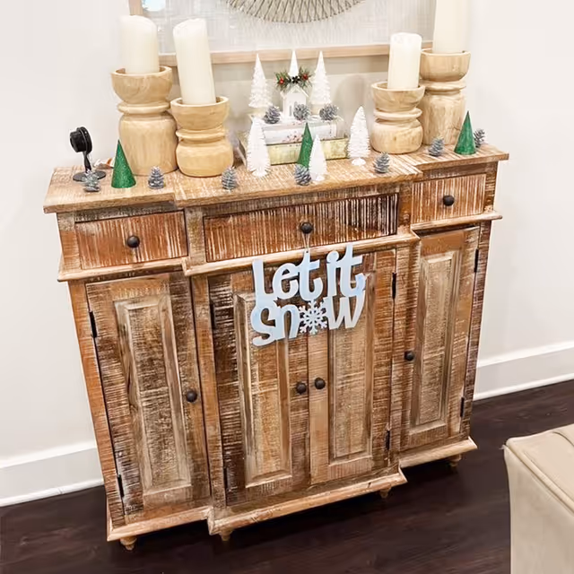 A rustic wooden cabinet decorated with small white and green Christmas trees, pine cones, and wooden candle holders with white candles. A decorative sign hanging on the cabinet reads 'Let it Snow'. The cabinet is placed against a white wall on a dark wooden floor.