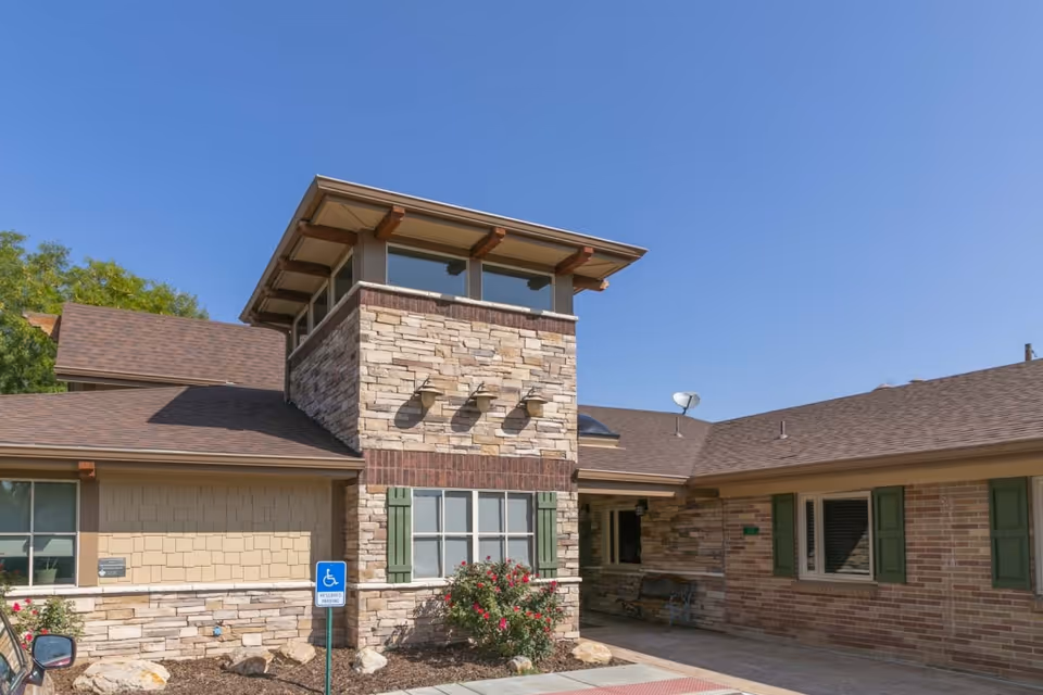 Front exterior of a single-story stone and brick care facility featuring a tower-like entrance, windows with green shutters, landscaping, and a handicap parking sign under a clear blue sky.