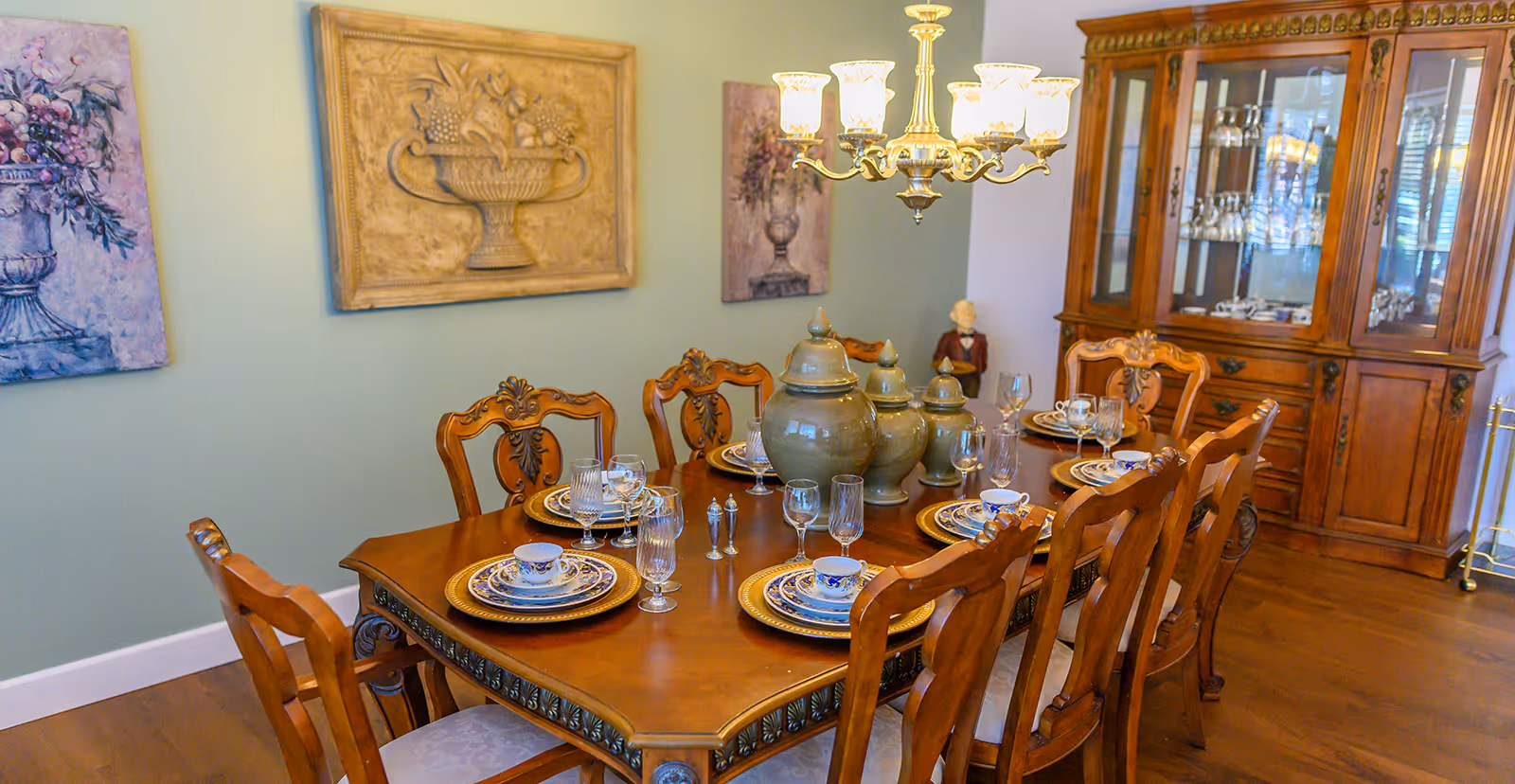 A formal dining room with a wooden dining table set for six, featuring ornate chairs, decorative plates, cups, and glassware. Three large ceramic jars are placed in the center of the table. The room has a wooden china cabinet filled with glassware and dishes, a chandelier with multiple lights hanging above the table, and artwork depicting floral arrangements on the walls.