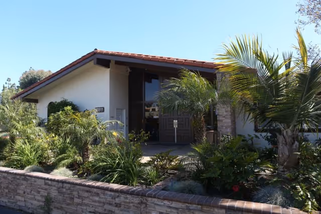 Exterior view of a single-story building with a tiled roof and large double doors, surrounded by lush green palm trees and other plants, with a low stone wall in front.