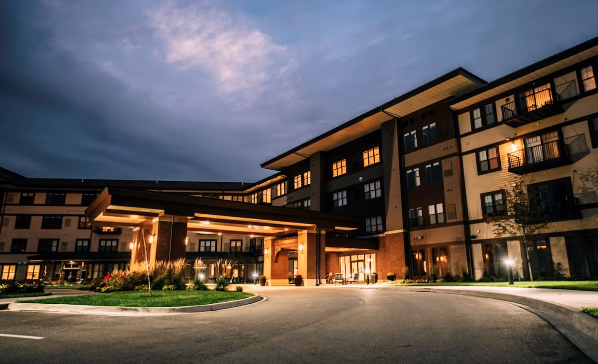 Exterior view of a multi-story senior living facility building at dusk with lights glowing from the windows and entrance area, featuring a covered driveway and landscaped greenery.