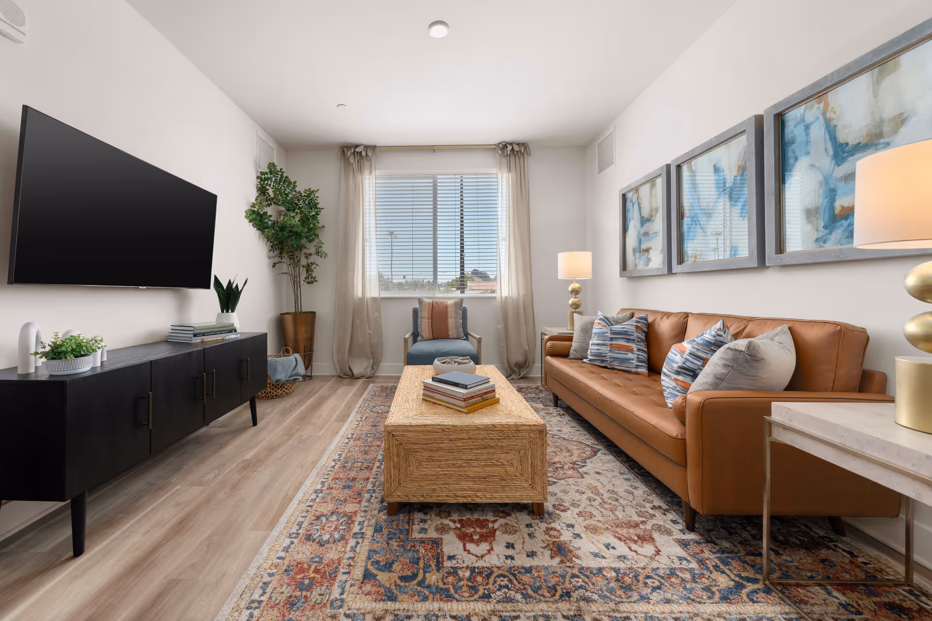Bright furnished living room with a leather sofa, patterned rug, coffee table, media console and wall-mounted TV under a large window.