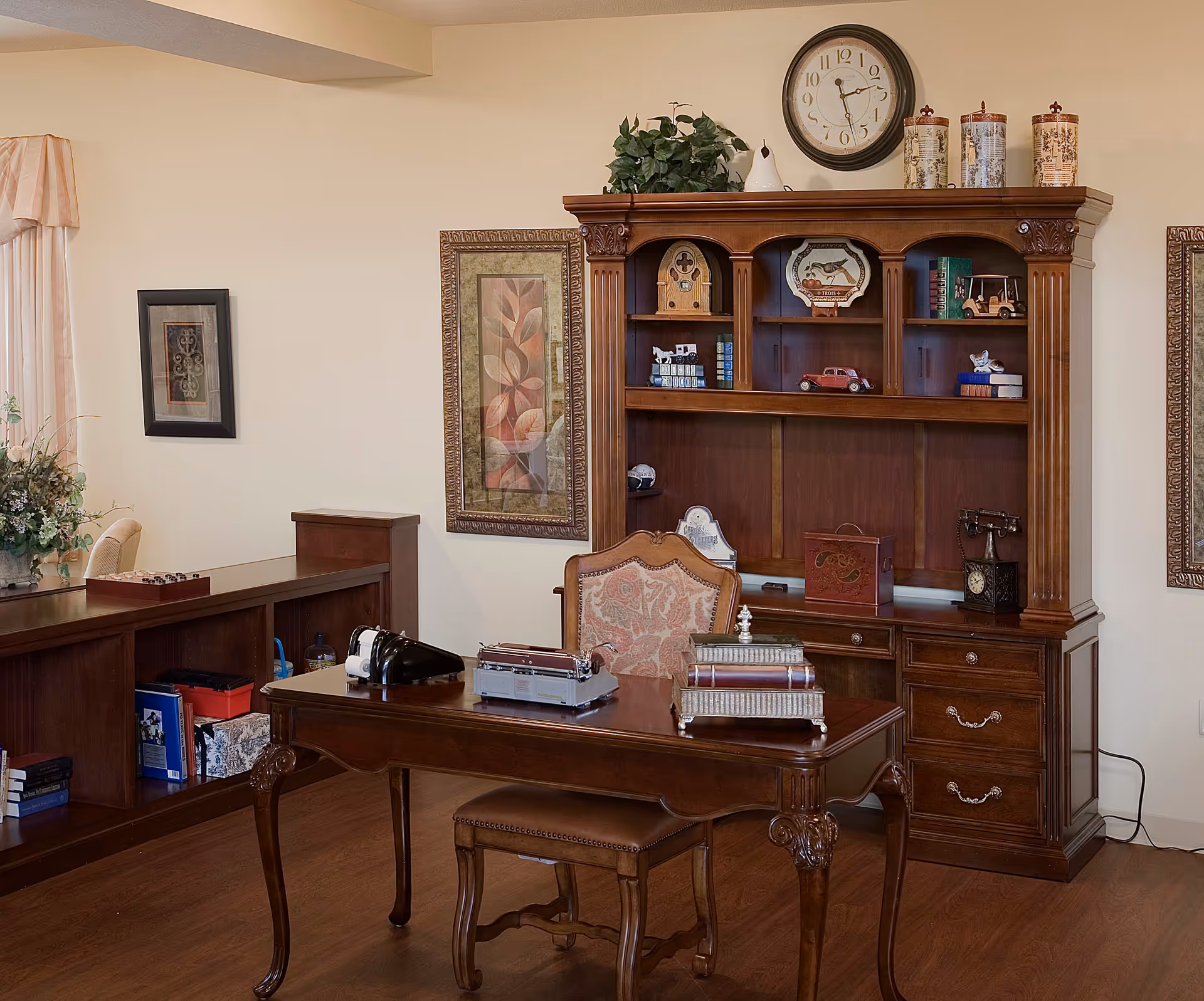 A traditional office or study room with a wooden desk and matching chair. The desk has decorative items including a typewriter, books, and a vintage telephone. Behind the desk is a large wooden hutch with shelves displaying various decorative objects, books, and a clock on top. The room has framed artwork on the walls and a wooden floor.