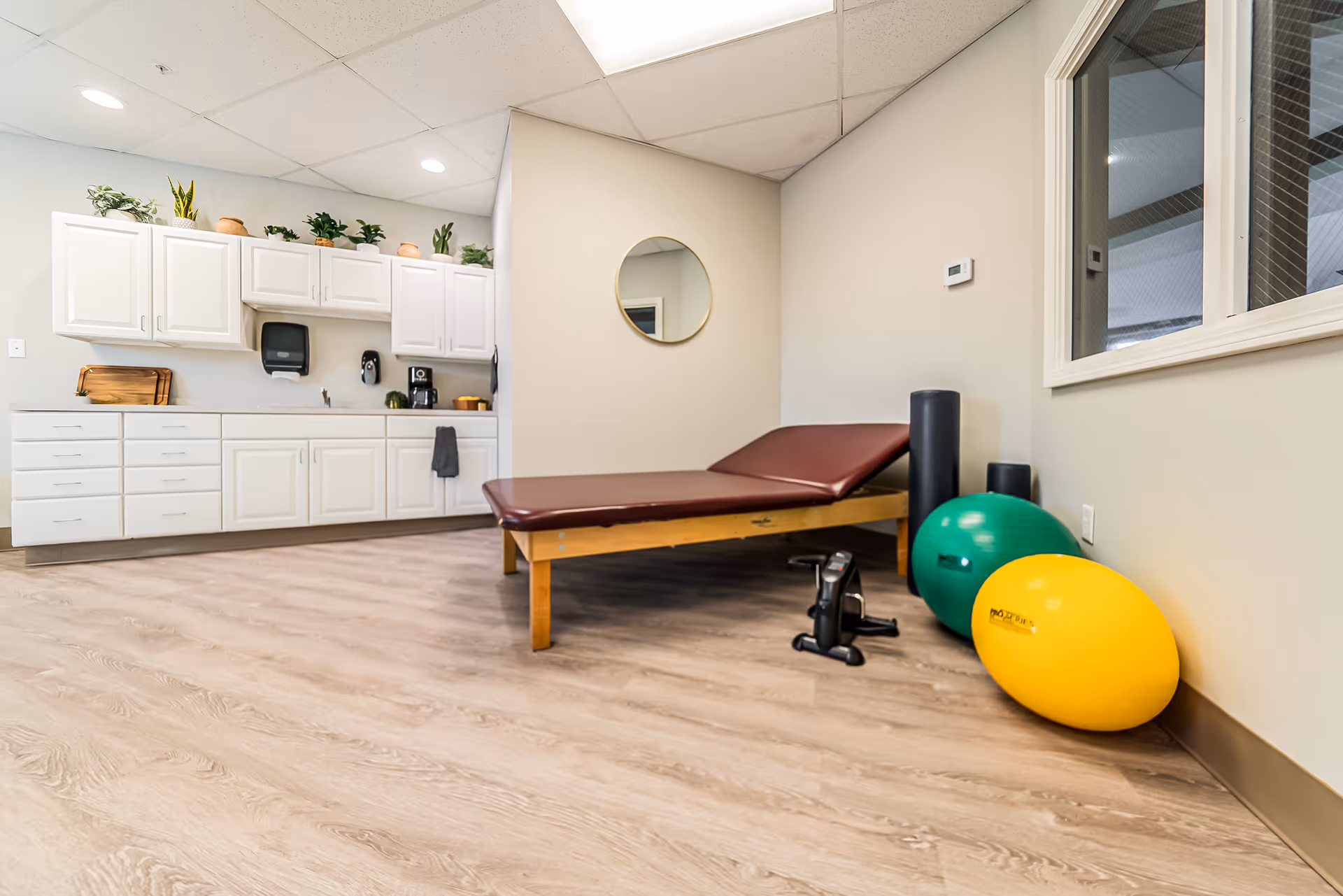 A therapy or exercise room with a maroon padded treatment table, two exercise balls (one green and one yellow), two black foam rollers, and a small pedal exerciser on a light wood floor. The room has white cabinets with plants on top, a paper towel dispenser, and a coffee maker. A round mirror hangs on the beige wall, and there is a window with a view into another room.