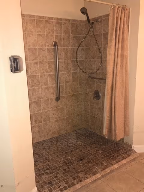 A tiled walk-in shower with a handheld showerhead attached to a flexible hose, a horizontal grab bar on the wall, and a beige shower curtain partially drawn to the side.