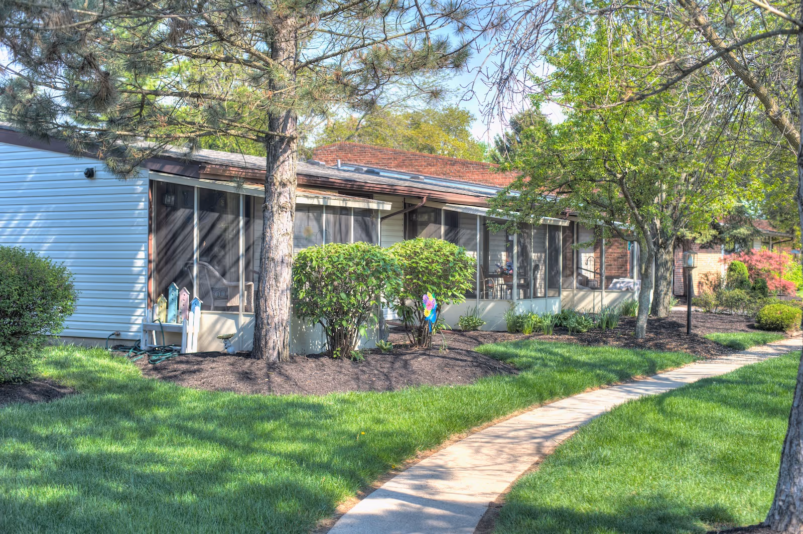 A sunny outdoor view of a single-story assisted living facility building with screened-in porches, surrounded by green grass, trees, and shrubs. A curved concrete walkway leads through the landscaped area in front of the building.