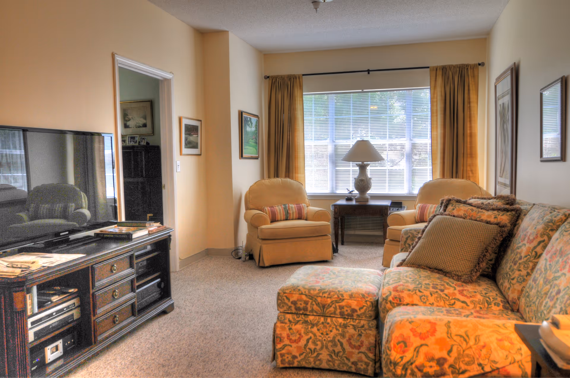 Sunlit living room with a floral sofa and ottoman, two armchairs by a large window, a lamp on a side table, and a TV on a wooden stand.
