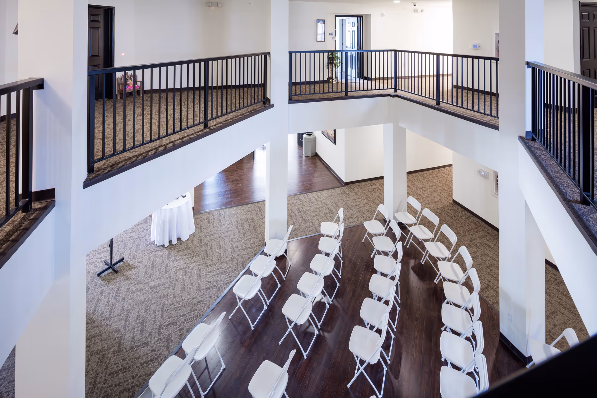 Interior view of a two-story common area in Newton Place Apartments with white folding chairs arranged in rows on a wooden floor below and carpeted walkways with black railings on the upper level.