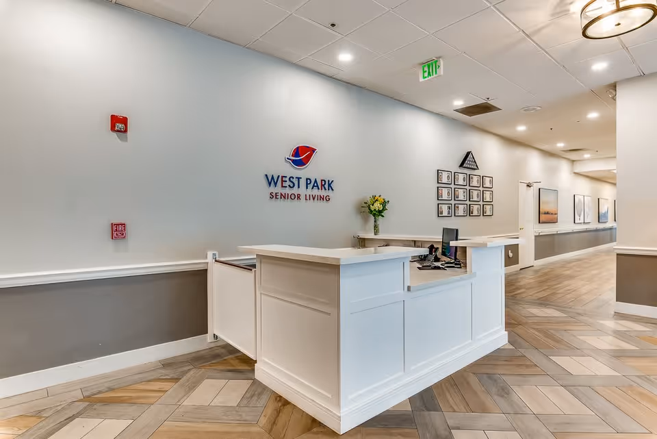 Reception desk area inside West Park Senior Living facility with a white counter, a computer, a vase with flowers, and a wall displaying the West Park Senior Living logo. The hallway extends to the right with framed pictures on the walls and a patterned wooden floor.