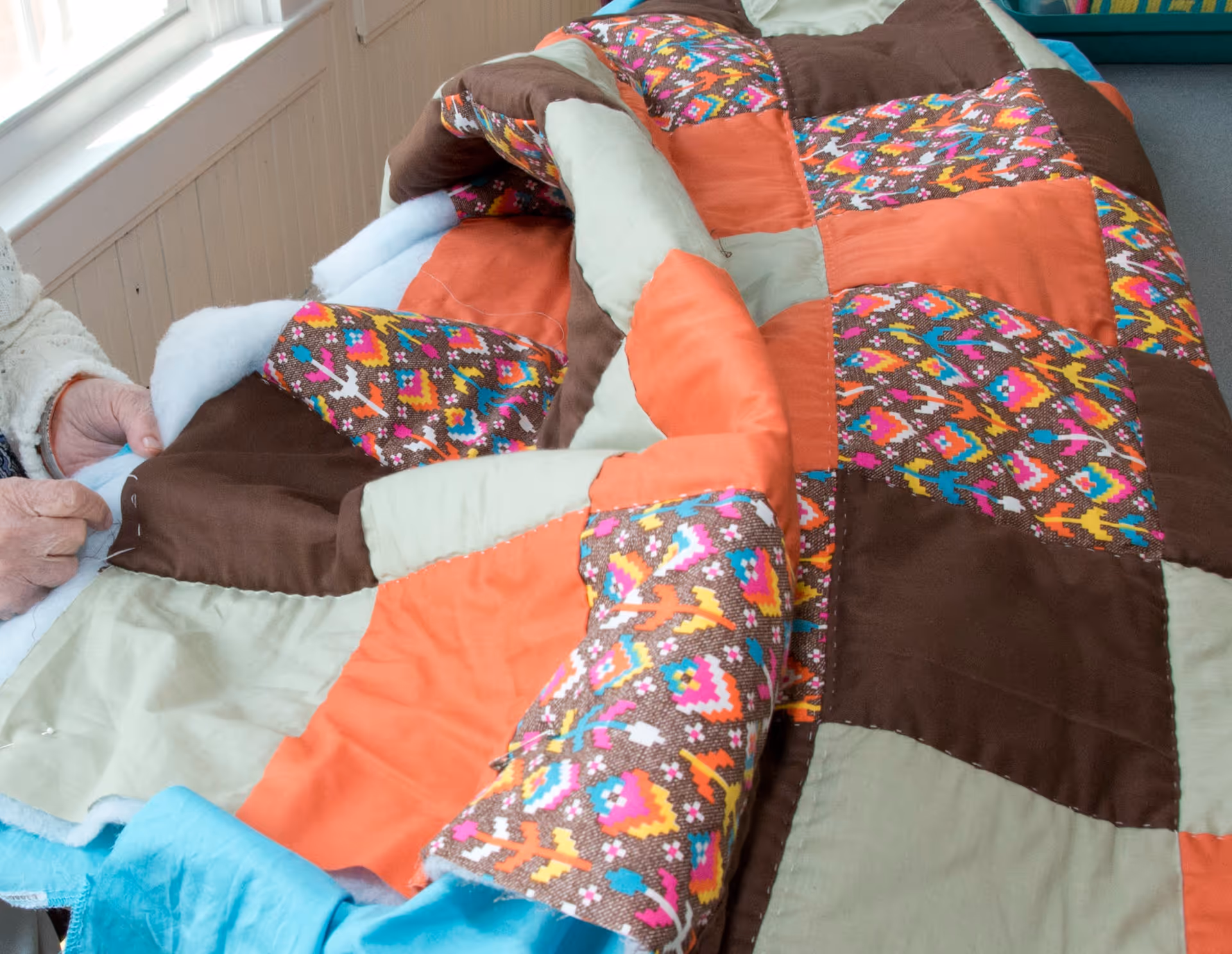 Close-up of a person sewing a colorful patchwork quilt with orange, brown, beige, and multicolored patterned fabric pieces.