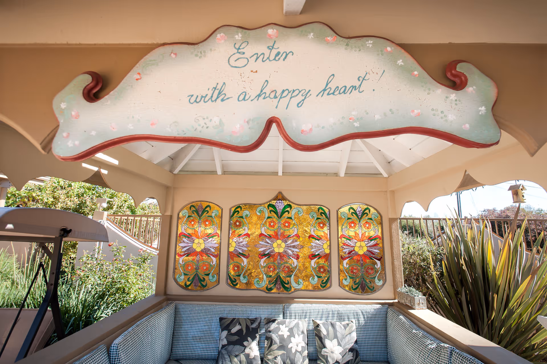 Covered outdoor seating area with a decorative sign reading "Enter with a happy heart", colorful stained-glass panels, and a cushioned bench.