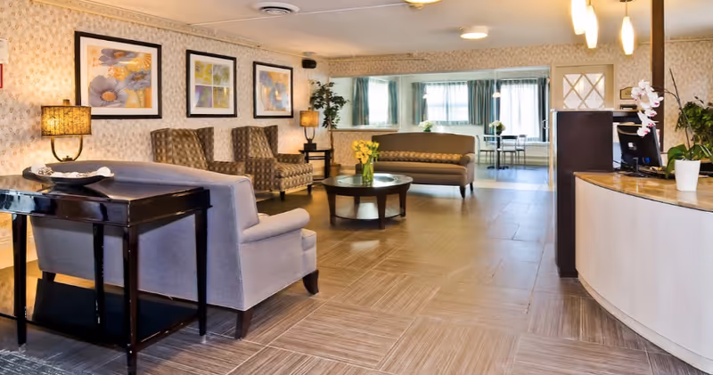 A spacious and well-lit living room area in a senior living facility with comfortable seating including armchairs and sofas, a round coffee table with a vase of yellow flowers, framed floral artwork on the walls, two table lamps, and a reception desk with a computer and potted plants.