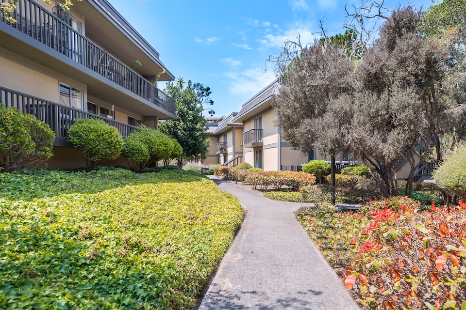 Outdoor pathway surrounded by well-maintained bushes and trees, leading between two beige multi-story residential buildings with balconies under a blue sky with some clouds.