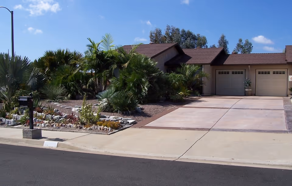 Exterior view of a single-story residential building with a double garage, a concrete driveway, and a landscaped front yard featuring various palm trees and other plants under a clear blue sky.