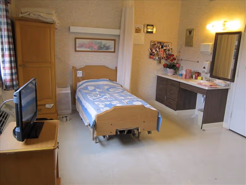 A single bed with a blue and white quilt in a senior care facility room. The room has a wooden wardrobe, a TV on a wooden stand, a window with plaid curtains, and a counter with a sink, mirror, and various personal items. The walls are decorated with a framed picture and a collage of photos.