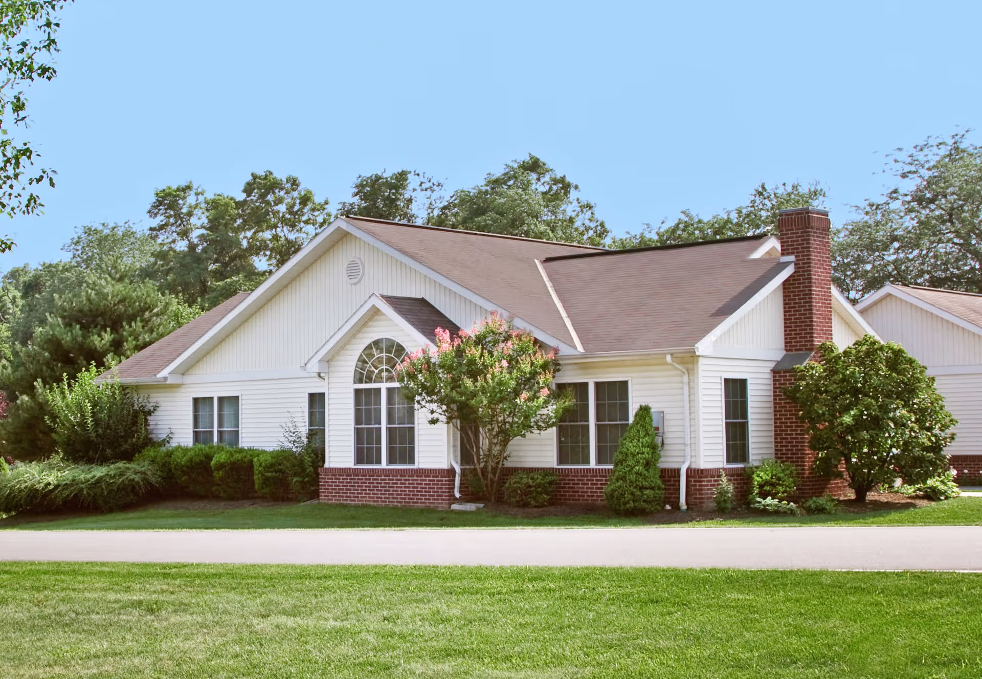 Exterior view of a single-story residential building with white siding and red brick accents, surrounded by green shrubs, trees, and a well-maintained lawn under a clear blue sky.