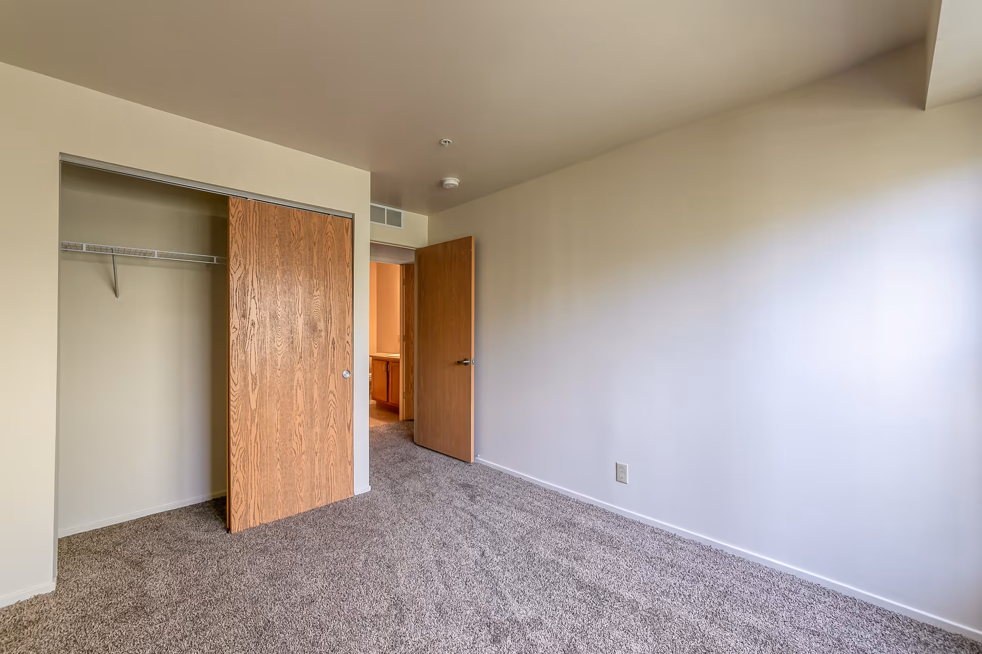 Empty bedroom with beige walls, carpeted floor, an open wooden closet door revealing a closet with a wire shelf, and an open wooden door leading to another room.
