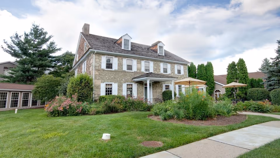 Stone-faced two-story building with a manicured lawn, flower beds, and patio umbrellas in front.