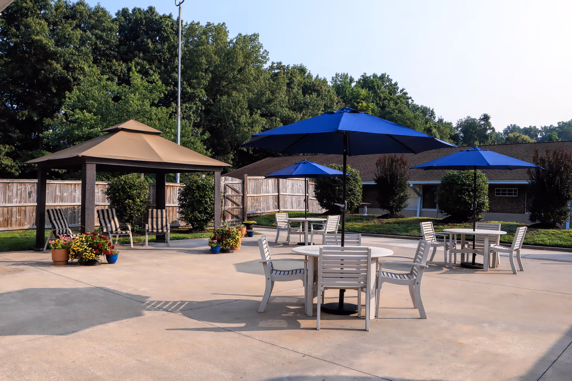 Outdoor patio area with round tables, chairs, blue umbrellas, and a gazebo near a low building.