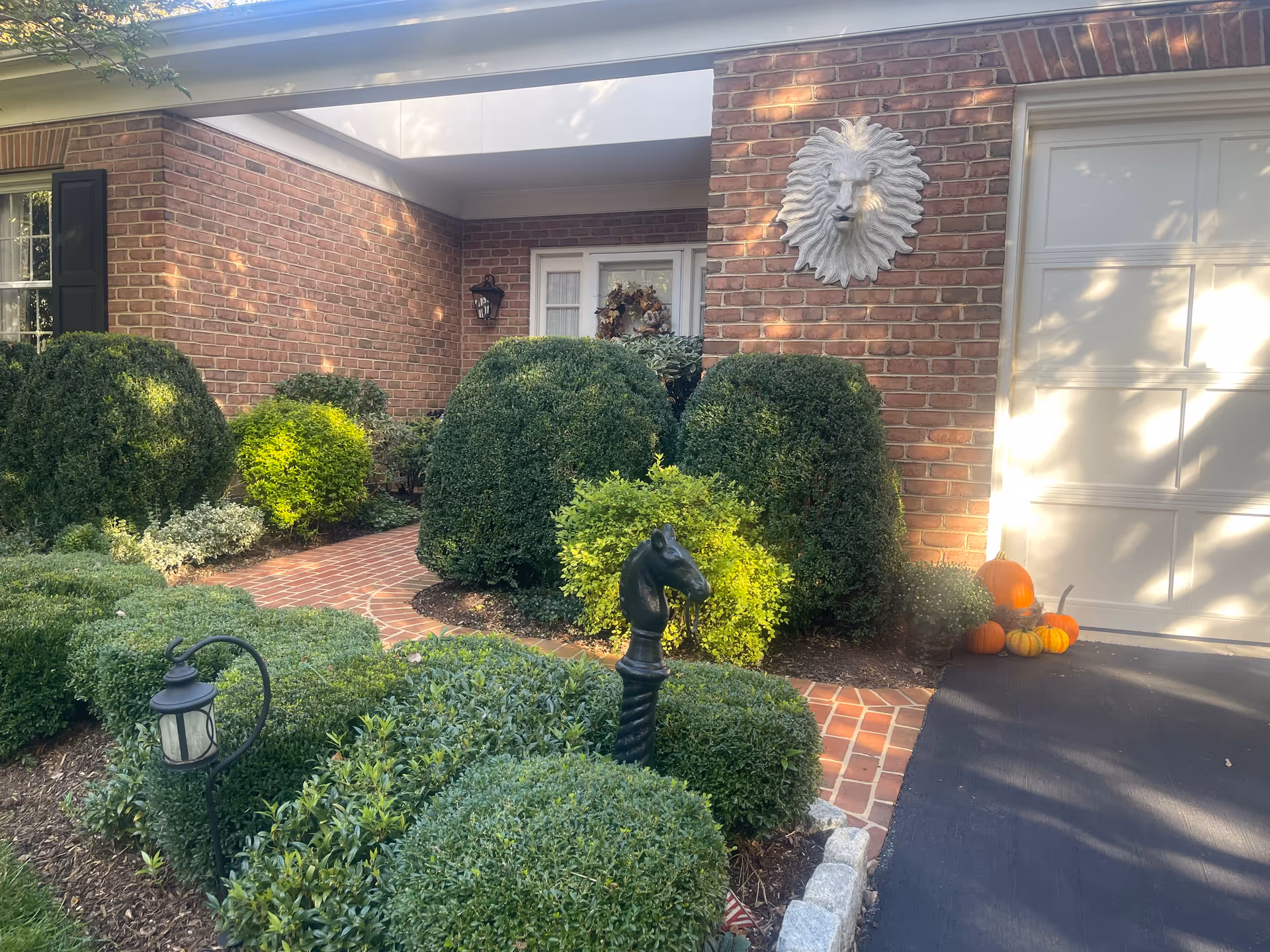 Front entrance of a brick building with neatly trimmed bushes and a brick walkway. There is a decorative lion head mounted on the wall near the garage door, and a small cluster of pumpkins placed on the driveway. A black horse head garden ornament and a small lantern are visible among the greenery.