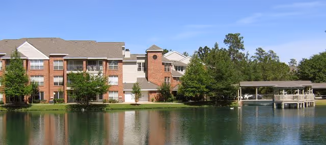 A multi-story brick and siding building beside a calm pond with trees and a covered wooden dock extending over the water under a clear blue sky.