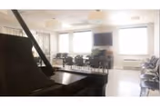 A bright room with large windows letting in natural light, featuring a grand piano in the foreground and rows of chairs arranged along the walls and in the center, with a flat-screen TV mounted on the wall and a plant beneath it.