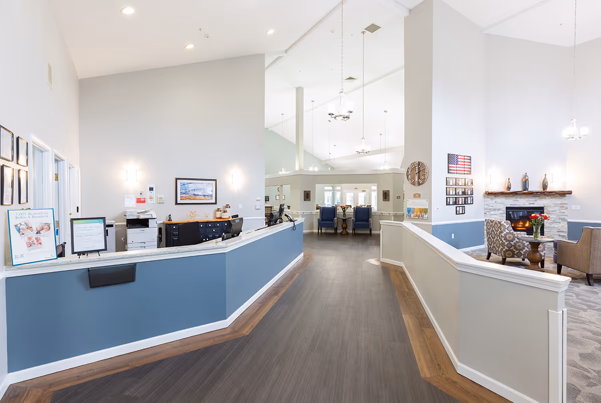 Interior view of a senior living facility reception area with a long blue and white front desk on the left, a seating area with chairs and a fireplace on the right, and a hallway leading to more seating and rooms in the background. The space is well-lit with high ceilings and modern light fixtures.