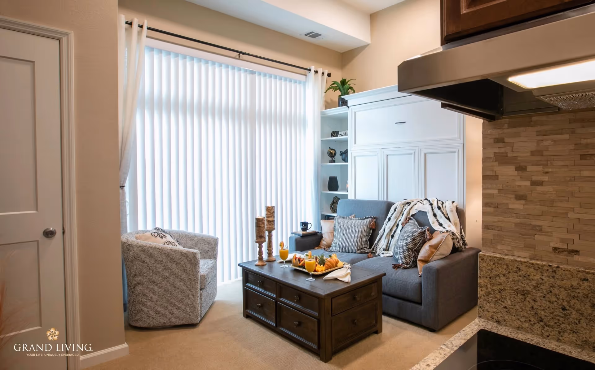 A cozy living room area with a gray sofa adorned with pillows and a throw blanket, a matching armchair, and a dark wooden coffee table holding candles, drinks, and snacks. The room has a large window with vertical blinds and light-colored walls. Part of a kitchen counter with granite countertop and a tiled backsplash is visible on the right side.