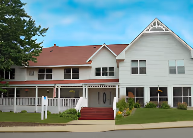 Front exterior view of a two-story white building with a red roof, featuring multiple windows and a covered porch with white railings. There is a small set of red stairs leading to the main entrance, surrounded by green grass and some shrubs. The sky is clear and blue.