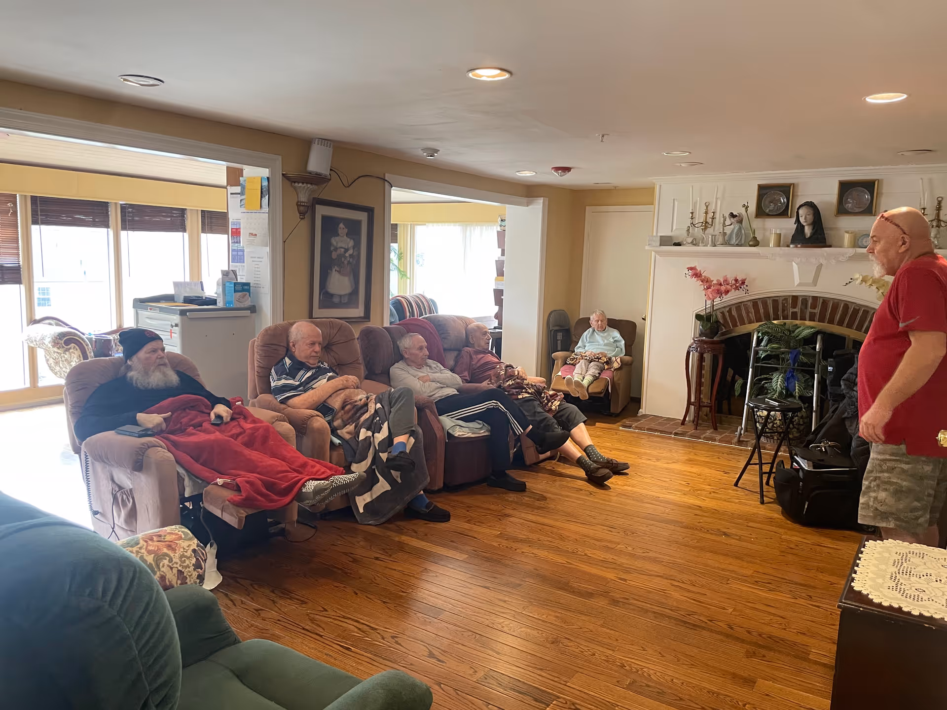 A group of elderly people sitting in recliner chairs in a cozy living room with wooden floors and a fireplace. One man stands on the right side of the room, engaging with the seated individuals. The room is warmly lit with natural light coming through large windows in the background.