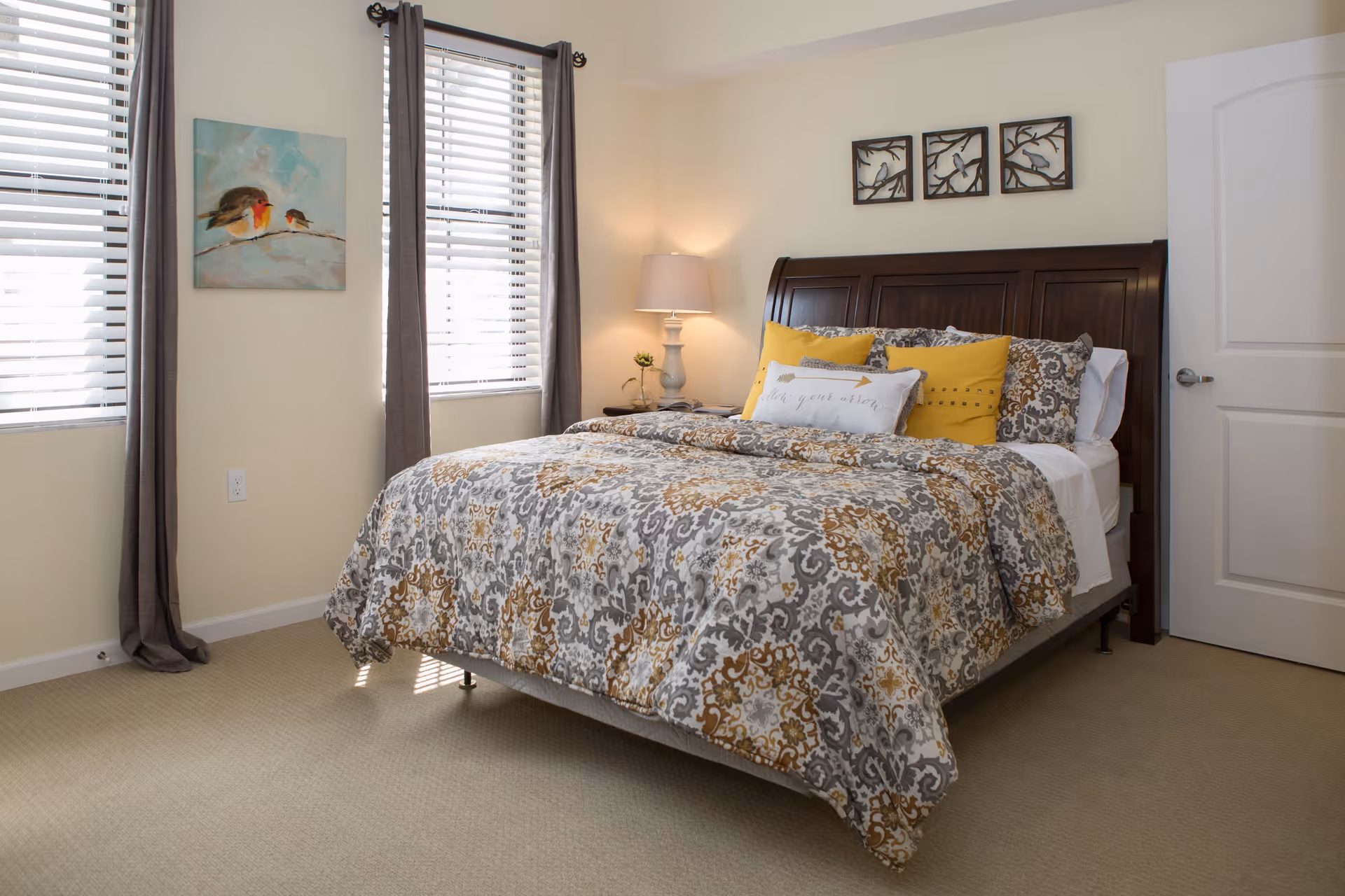 A cozy bedroom with a large bed featuring a patterned comforter in gray, white, and mustard colors. The bed has several pillows, including two mustard yellow ones and a decorative pillow with the text 'follow your arrow'. There is a wooden headboard and a nightstand with a lamp and a small plant. The room has beige walls, two windows with white blinds and gray curtains, and a door with a silver handle. Artwork featuring birds is hung on the walls.