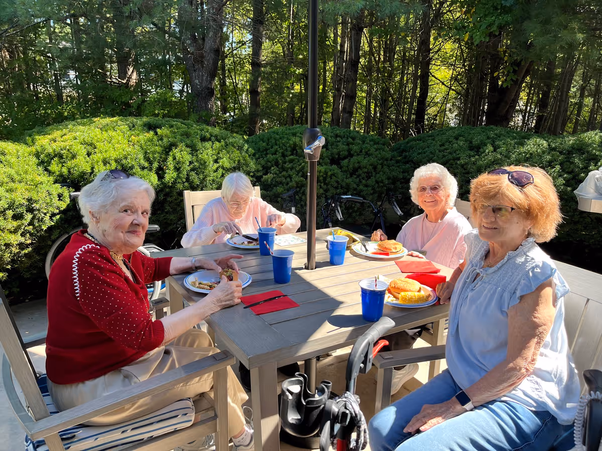 Four elderly women sitting around a wooden outdoor table enjoying a meal together. The table has plates with food, blue cups, and red napkins. The setting is surrounded by green bushes and trees, with sunlight filtering through.