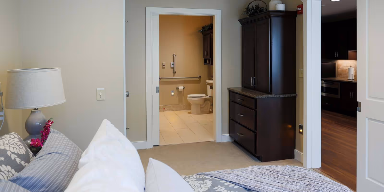 Furnished bedroom showing a bed with pillows and lamp in the foreground, a dark wood dresser to the right, and open doorways to a bathroom and kitchen beyond.