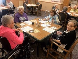Four elderly women sitting around a rectangular dining table in a communal dining area, engaging in conversation and activities. The table has cups, plates, and papers on it. Other tables and chairs are visible in the background with additional elderly residents seated.