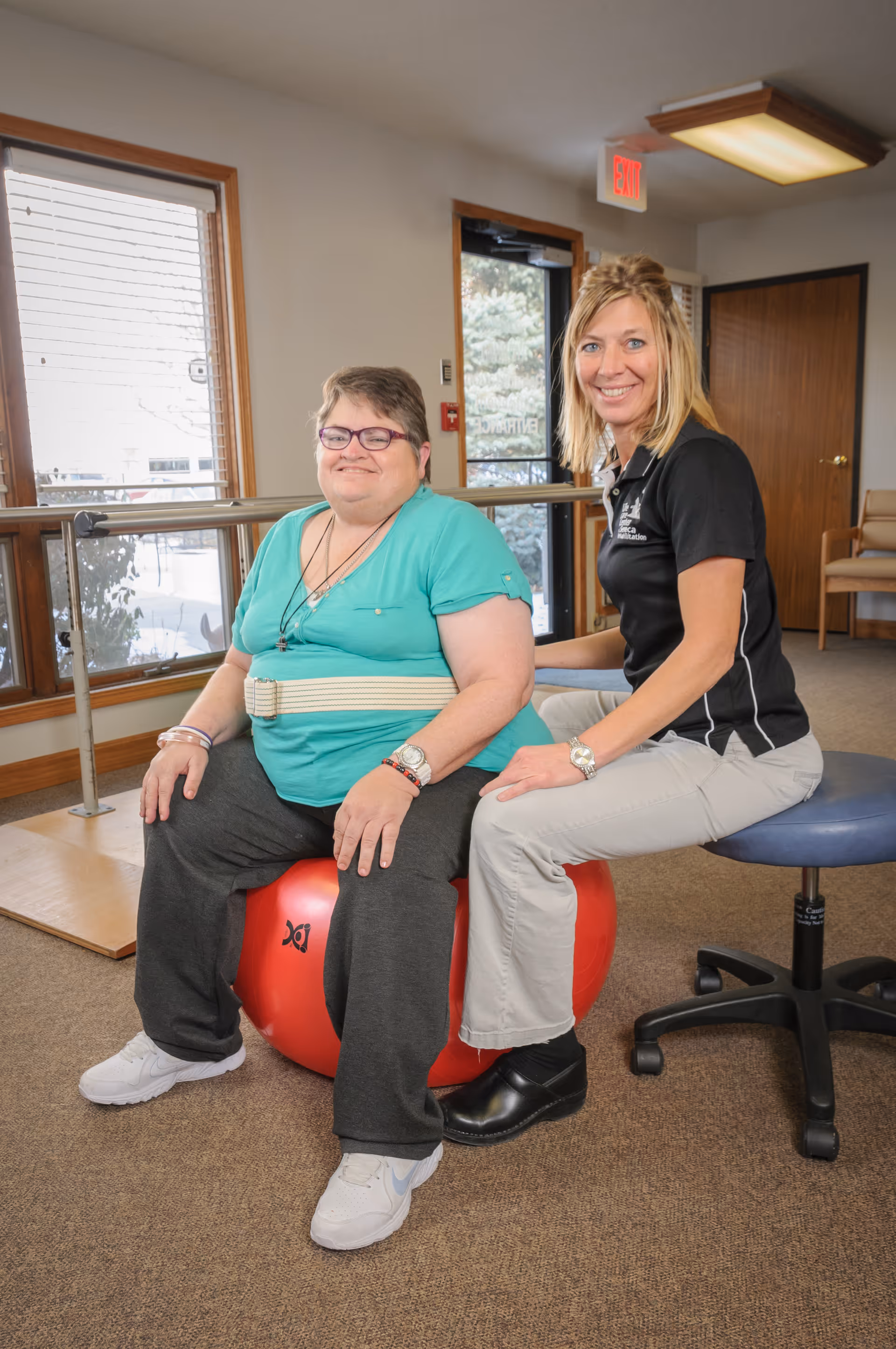 A woman in a teal shirt and dark pants sits on a red exercise ball with a supportive belt around her waist, smiling. Next to her, a woman in a black polo shirt and light-colored pants sits on a rolling stool, also smiling. They are in a room with large windows, a wooden door, and handrails along the wall.