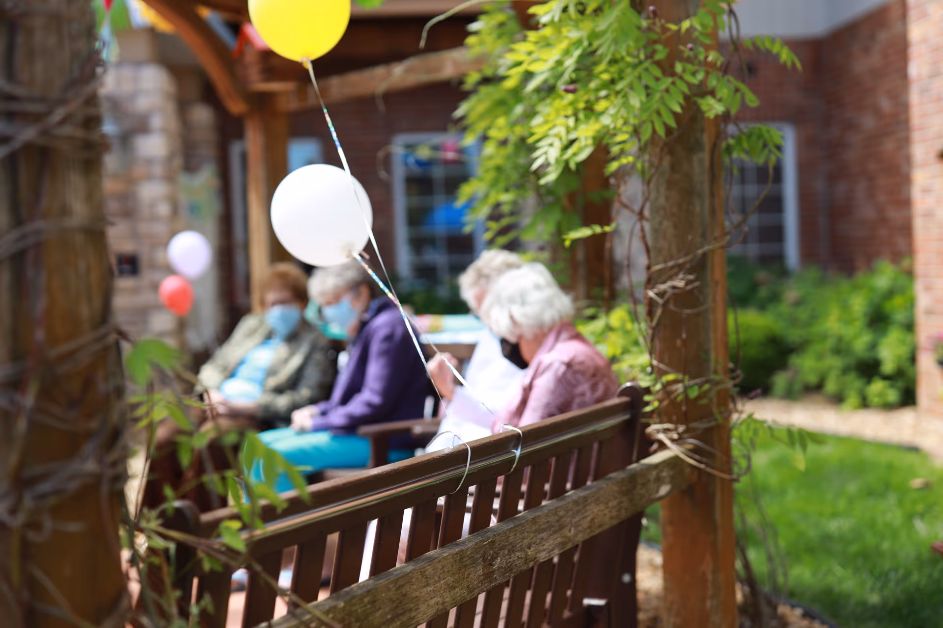 Several elderly residents wearing masks sit on wooden benches in a garden under a pergola decorated with colorful balloons.