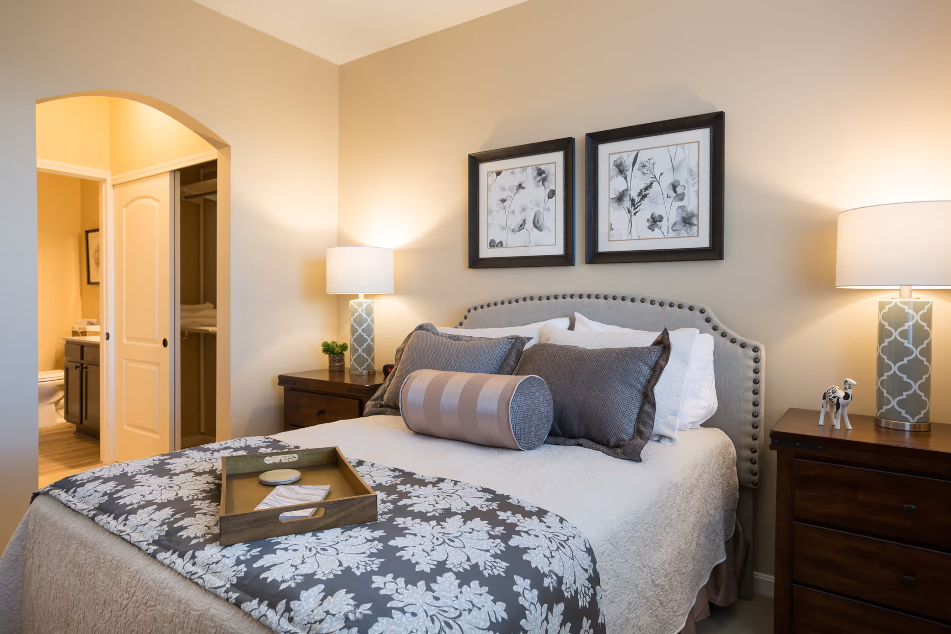 A cozy bedroom with a neatly made bed featuring multiple pillows and a decorative bolster pillow. The bed has a patterned gray and white bedspread. On either side of the bed are wooden nightstands with matching table lamps. Above the bed are two framed black and white botanical prints. An open doorway reveals a glimpse of a bathroom with a toilet and vanity.