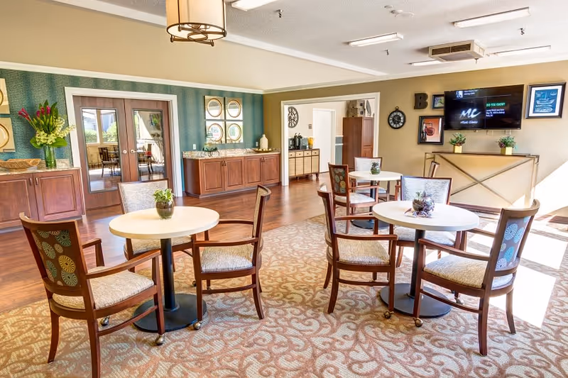 A bright and spacious senior living common area with round tables and wooden chairs arranged on a patterned carpet. The room features wooden cabinetry along the walls, decorative plants on the tables, a wall-mounted TV, and large windows allowing natural light to fill the space.